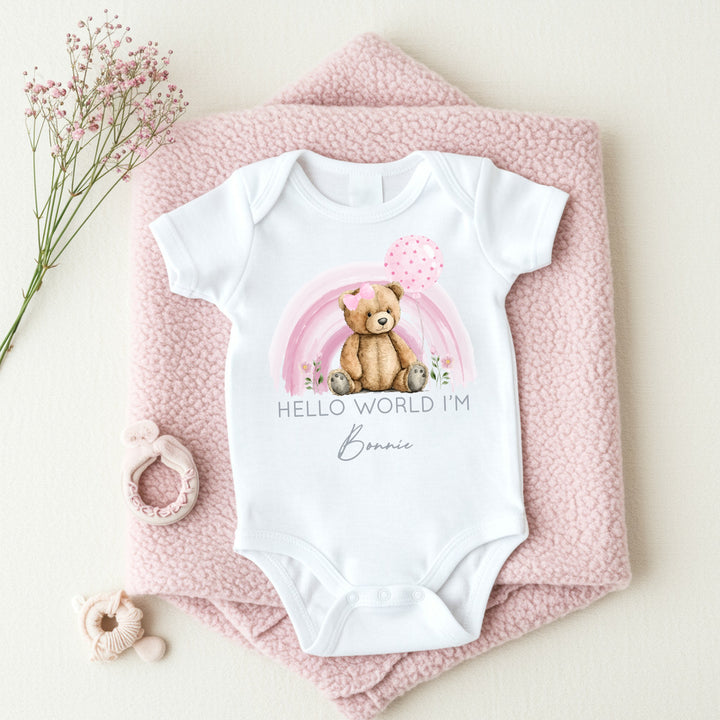 a personalised white baby vest that says 'Hello World I'm Bonnie'. This design features a pink rainbow with teddy infront of it 