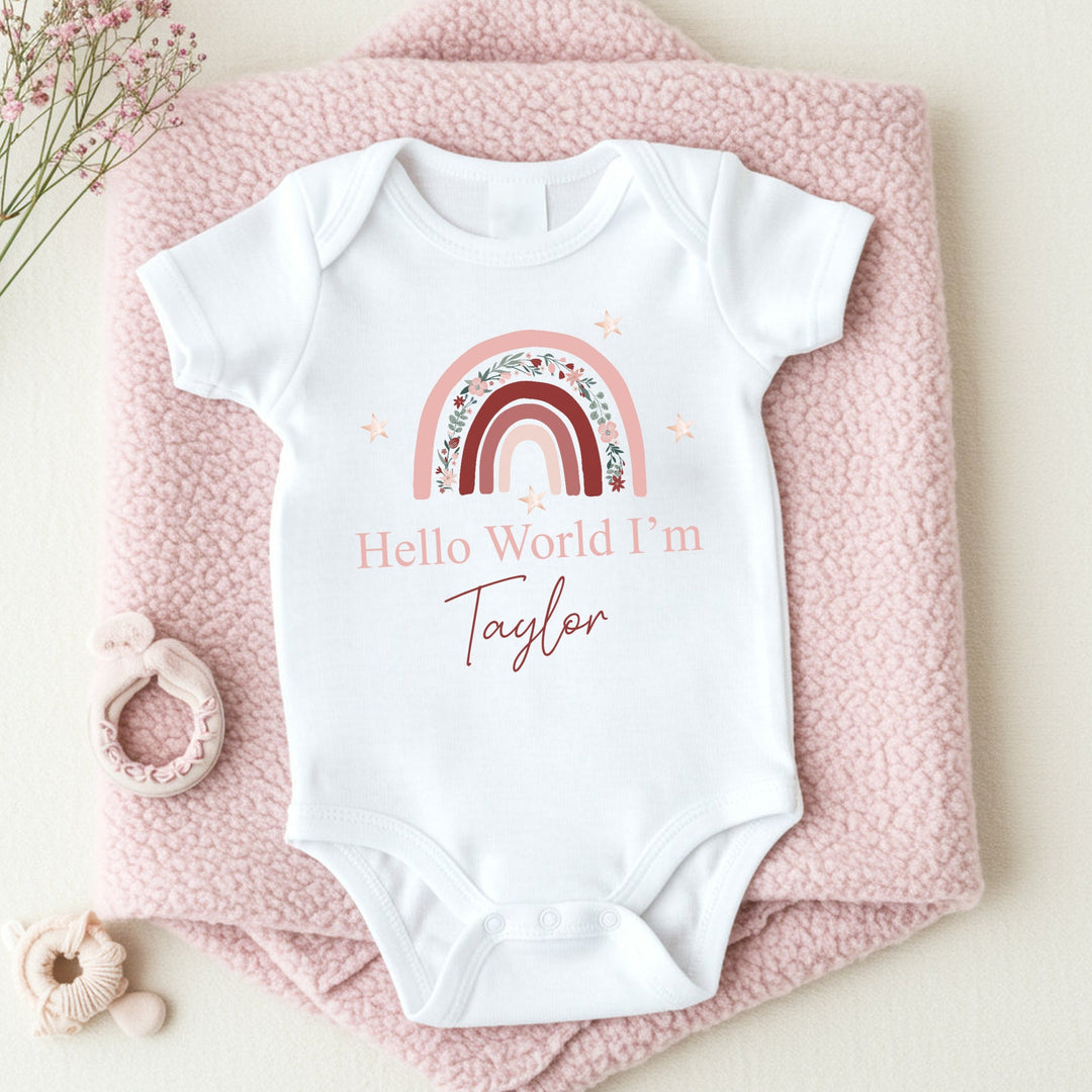 a personalised white baby vest that says 'Hello World I'm Taylor'. This design features a pink floral rainbow