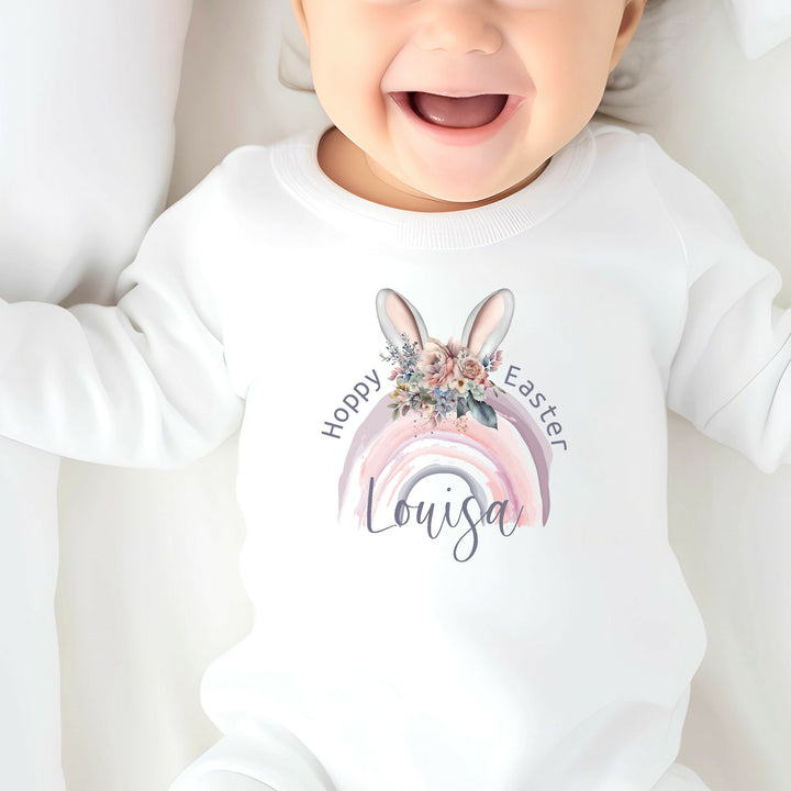 Personalised white Easter baby grow/sleepsuit that says 'Hoppy Easter Louisa'. This design features a purple rainbow with bunny ears on top