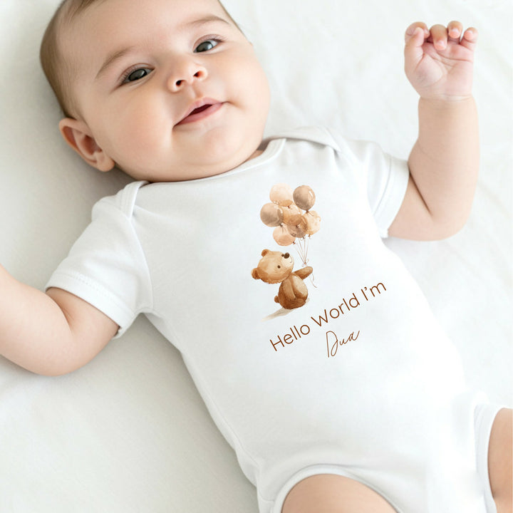 A personalised white baby vest that says 'Hello World I'm Dua'. This design features a little brown bear holding a bunch of balloons