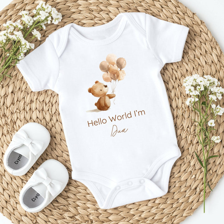 A personalised white baby vest that says 'Hello World I'm Dua'. This design features a little brown bear holding a bunch of balloons