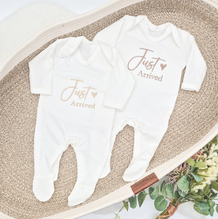 This photo shows 2 white babygrows that have 'Just Arrived' embroidered on them. This design features a small heart and shows 2 of the avaliable font colours (Sand and Fawn)