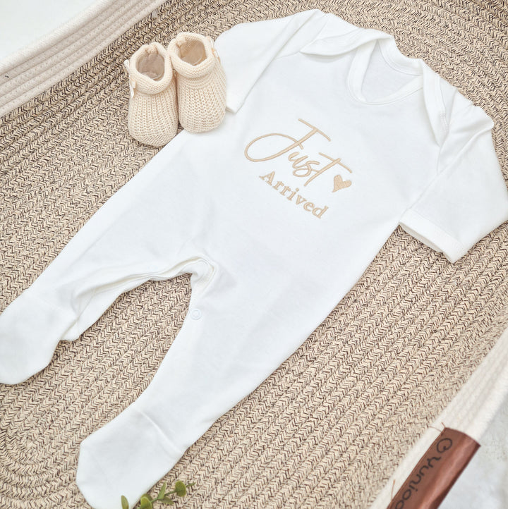 This photo shows a white babygrows that have 'Just Arrived' embroidered on them. This design features a small heart and shows one of the avaliable font colours (Sand)
