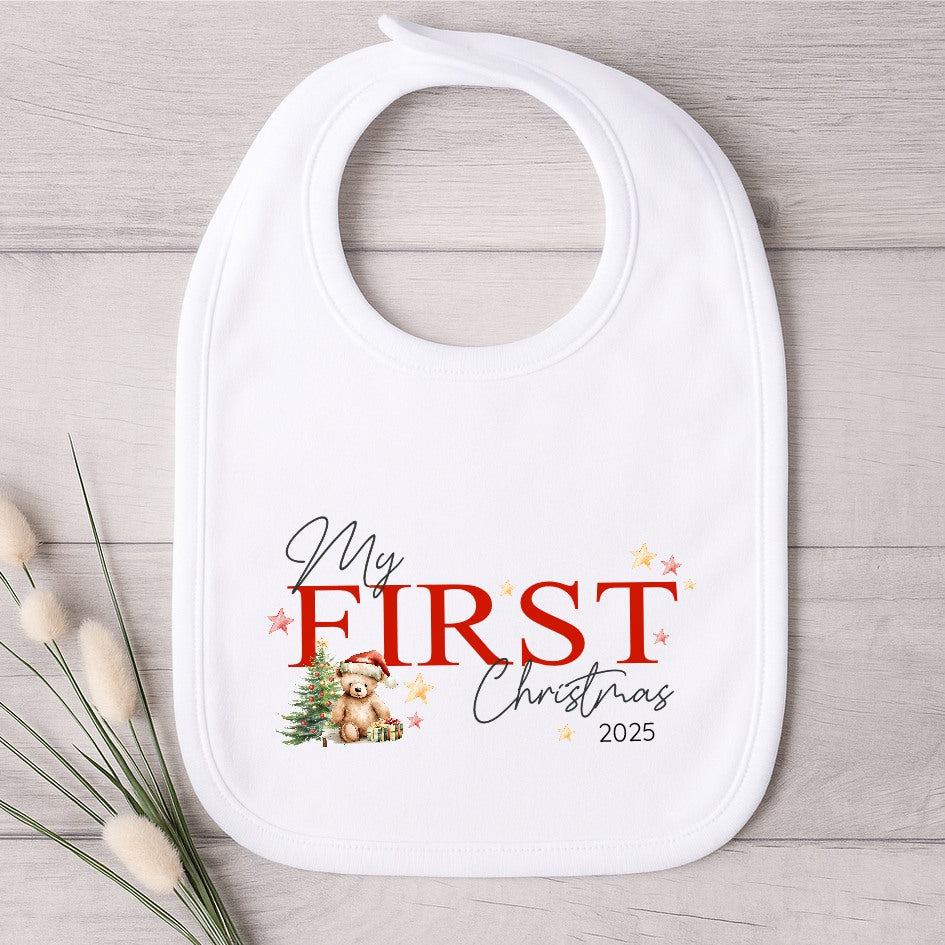 White baby bib with 'My First Christmas 2025' text and bear illustration on a wooden surface.