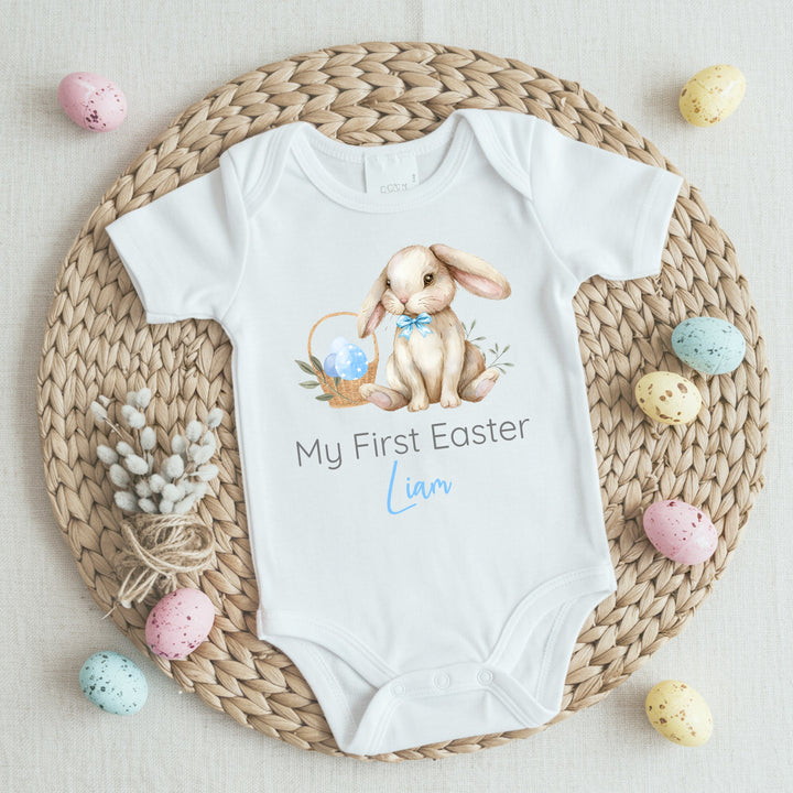 Personalised white Easter baby vest that says 'My First Easter Liam'. This design features a brown rabbit wearing a blue bow tie sitting next to a basket filled with Easter eggs