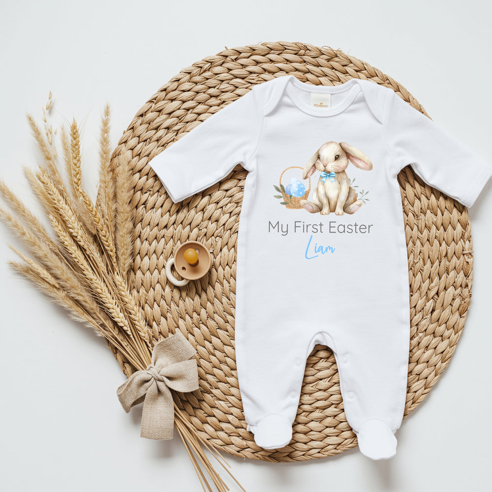 Personalised white Easter baby grow/sleepsuit that says 'My First Easter Liam'. This design features a brown rabbit wearing a blue bow tie sitting next to a basket filled with Easter eggs