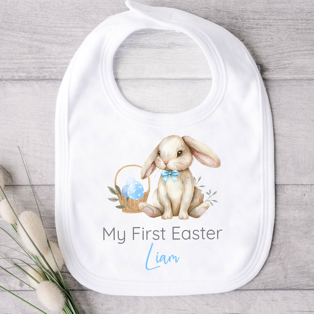 Personalised white Easter baby bib that says 'My First Easter Liam'. This design features a brown rabbit wearing a blue bow tie sitting next to a basket filled with Easter eggs