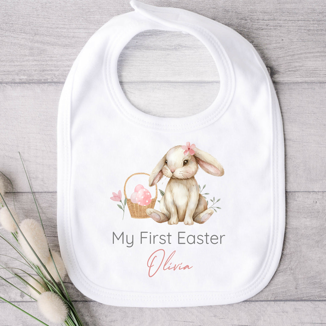 Personalised white Easter baby bib that says 'My First Easter Olivia'. This design features a brown bunny wearing a pink bow sitting next to a basket with pink eggs in it