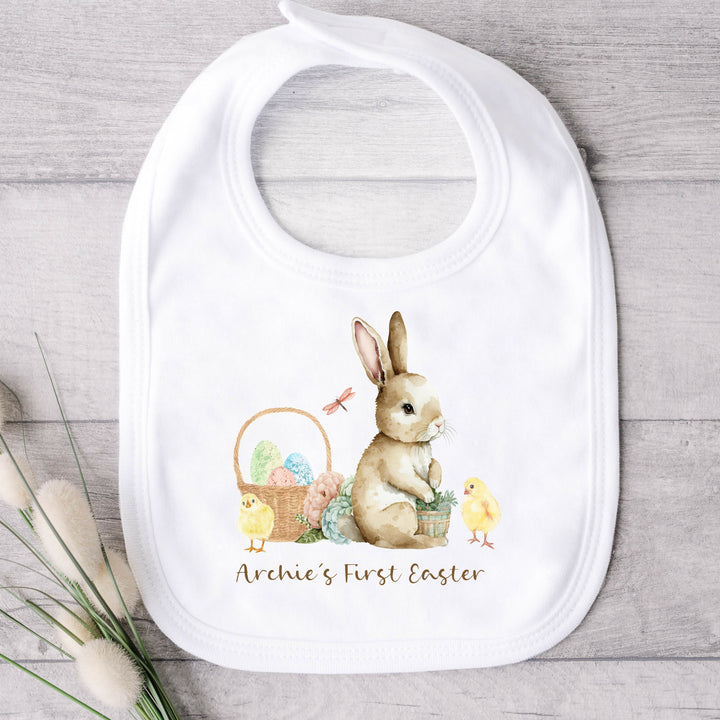 Personalised white Easter baby bib that says 'Archie's First Easter'. This design features a brown rabbit sitting next to an Easter basket and 2 yellow chicks