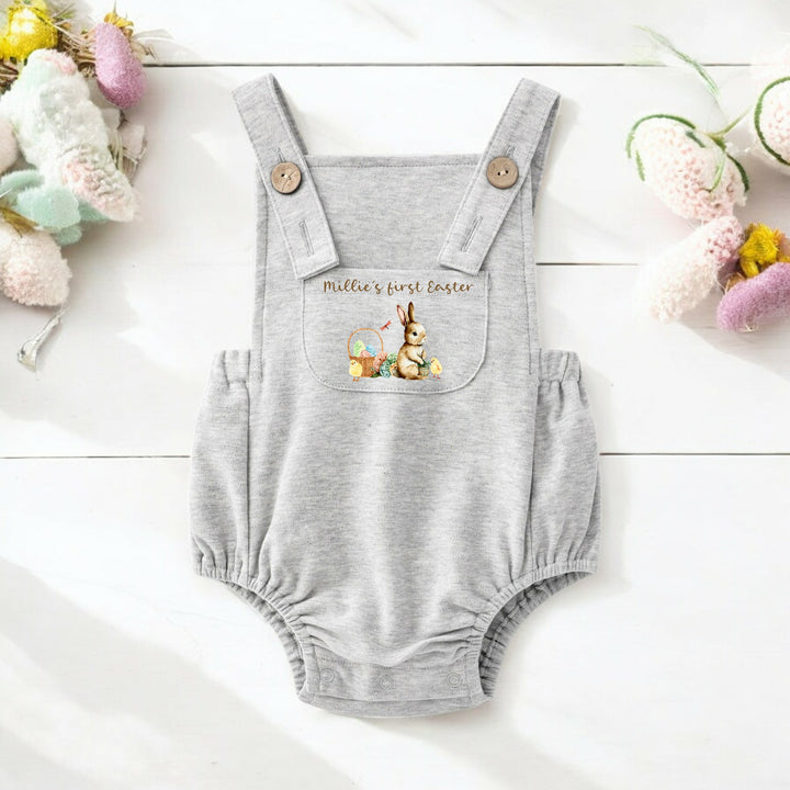 Personalised grey Easter dungarees that say 'Millie's First Easter'. This design features a brown bunny sittinhg next to an Easter basket full of eggs