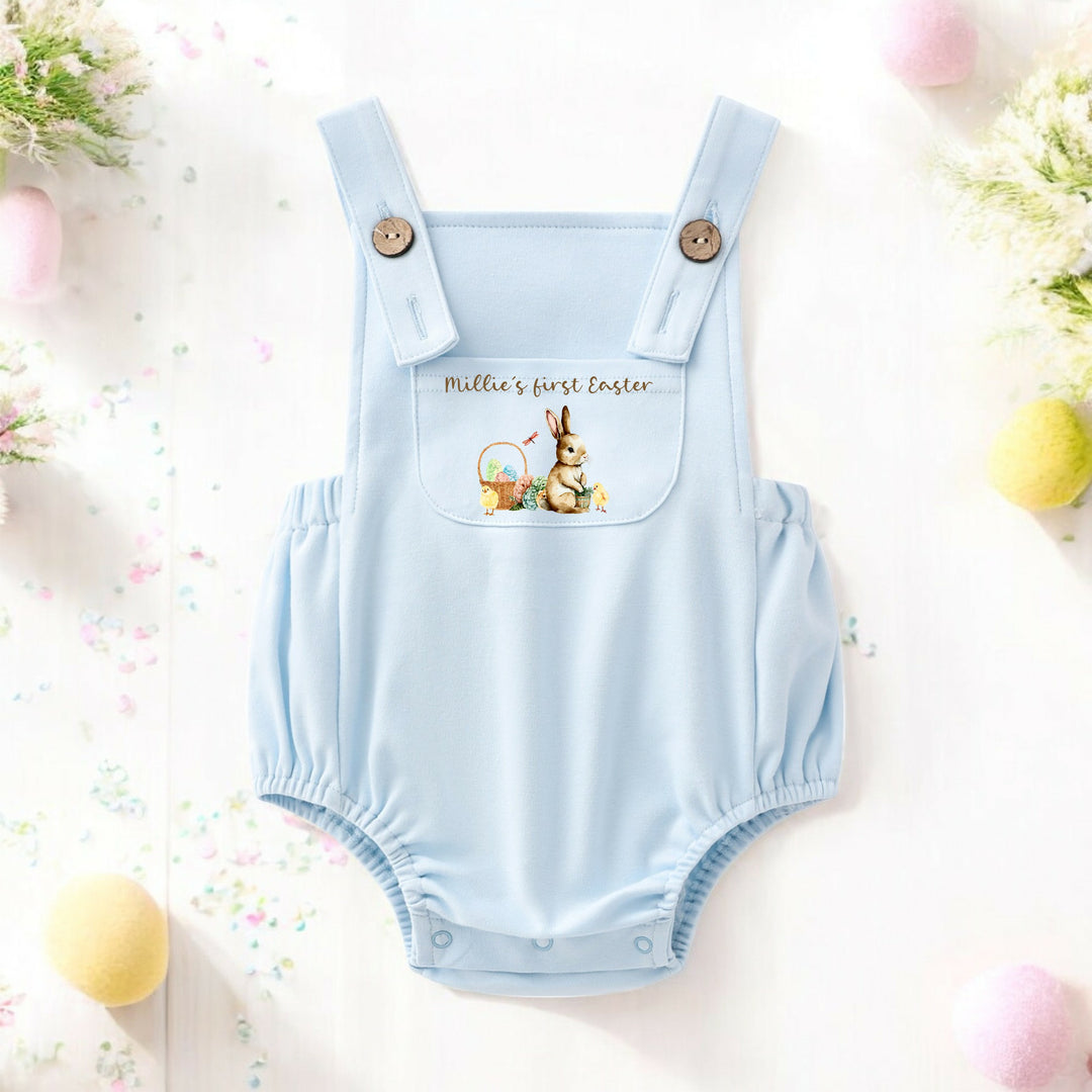 Personalised blue Easter dungarees that say 'Millie's First Easter'. This design features a brown bunny sittinhg next to an Easter basket full of eggs