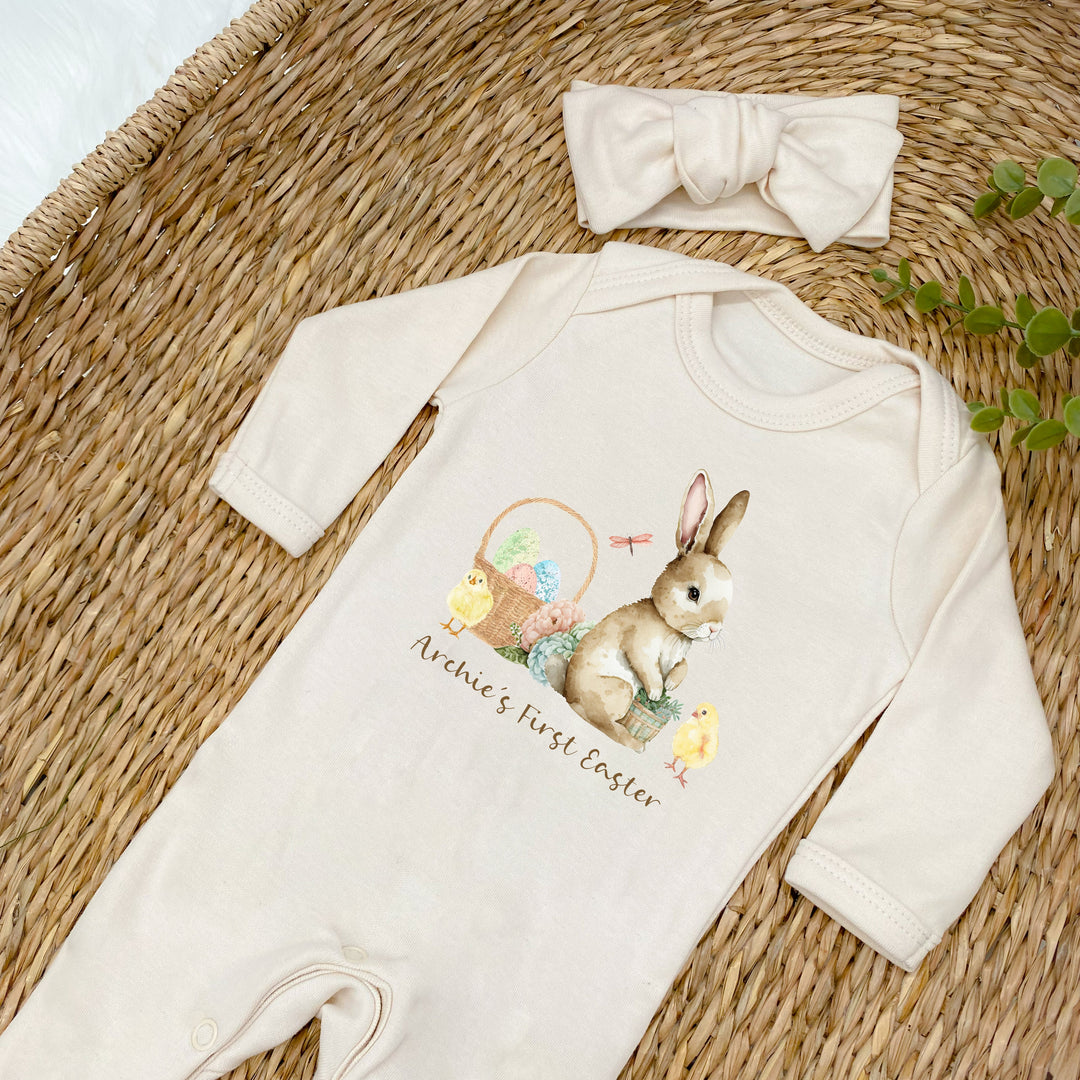 Personalised beige Easter baby grow/sleepsuit that says 'Archie's First Easter'. This design features a brown rabbit sitting next to an Easter basket and 2 yellow chicks