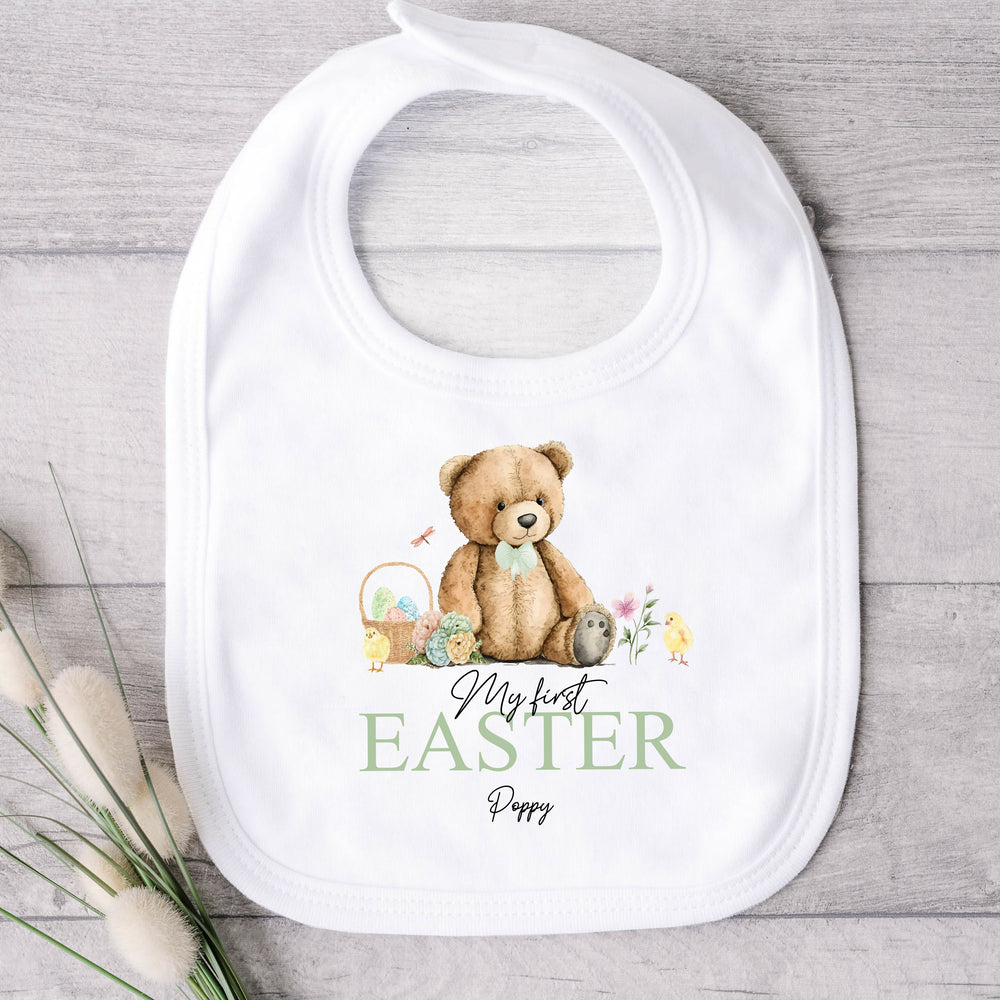 Personalised white Easter baby bib that says 'My First Easter Wallace'. This design features a teddy bear wearing a sage green bowtie sitting next to an Easter basket and 2 yellow chicks