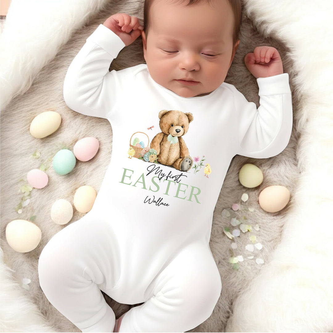 Personalised white Easter baby grow/sleepsuit that says 'My First Easter Wallace'. This design features a teddy bear wearing a sage green bowtie sitting next to an Easter basket and 2 yellow chicks