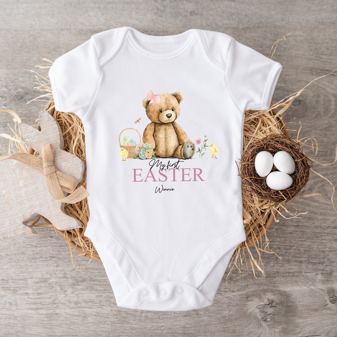 Personalised white Easter baby vest that says 'My First Easter Winnie'. This design features a teddy bear with a pink bow sitting next to an Easter basket filled with eggs and 2 yellow chicks