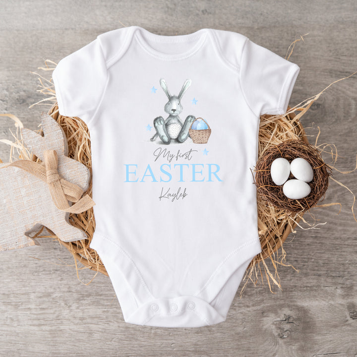 Personalised white Easter baby vest that says 'My First Easter Kayleb'. This design features a blue rabbit sitting next to an Easter basket full of blue eggs