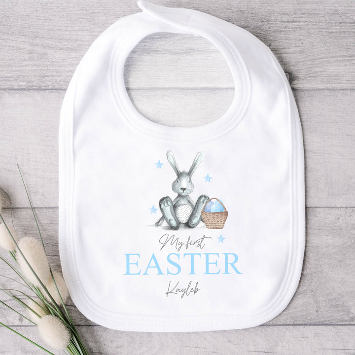 Personalised white Easter baby bib that says 'My First Easter Kayleb'. This design features a blue rabbit sitting next to an Easter basket full of blue eggs