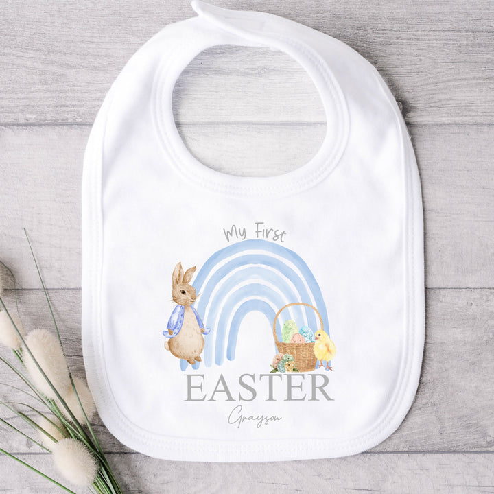 Personalised white Easter baby bib that says 'My First Easter Grayson'. This design features a blue rainbow with a rabbit wearing a blue jacket one side and an Easter basket and a chick the other