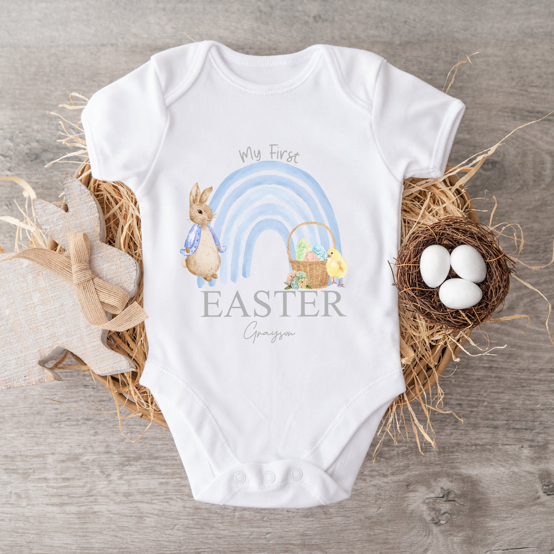 Personalised white Easter baby vest that says 'My First Easter Grayson'. This design features a blue rainbow with a rabbit wearing a blue jacket one side and an Easter basket and a chick the other