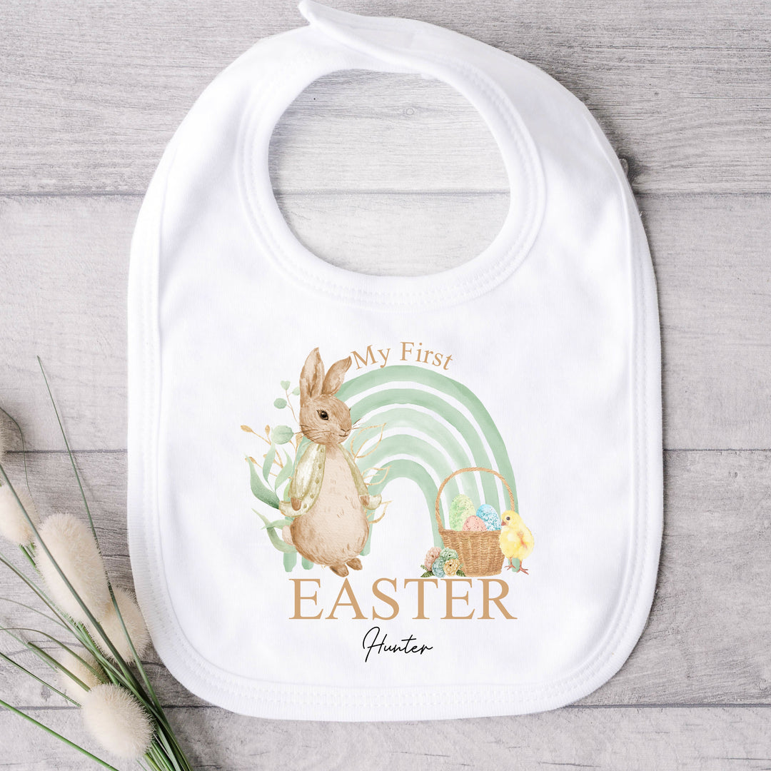 Personalised white Easter baby bib that says 'My First Easter Hunter'. This design features a sage green rainbow with a rabbit wearing a beige jacket one side and an Easter basket and a chick the other