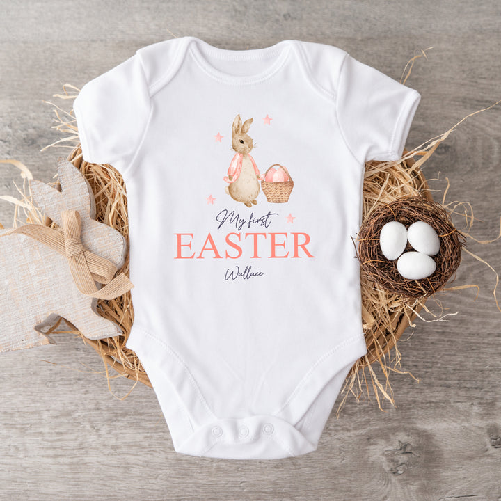 Personalised white Easter baby vest that says 'My First Easter Olivia'. This design features a rabbit wearing a pink jacket holding an Easter basket surrounded by pink stars