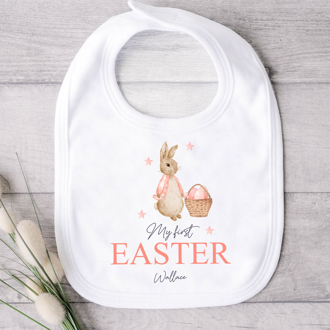 Personalised white Easter baby bib that says 'My First Easter Olivia'. This design features a rabbit wearing a pink jacket holding an Easter basket surrounded by pink stars