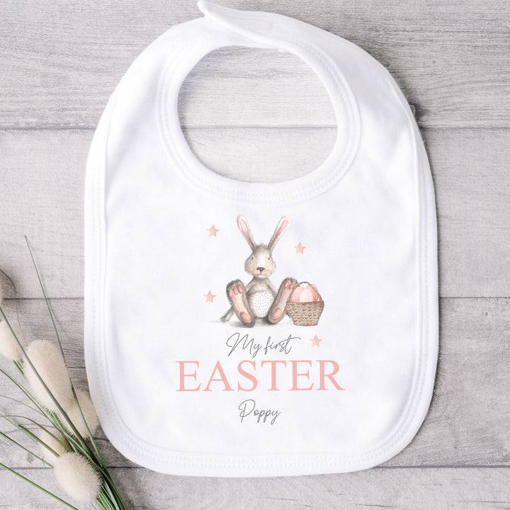 Personalised white Easter baby bib that says 'My First Easter Poppy'. This design features a grey rabbit with pink details sitting next to an Easter basket full with eggs