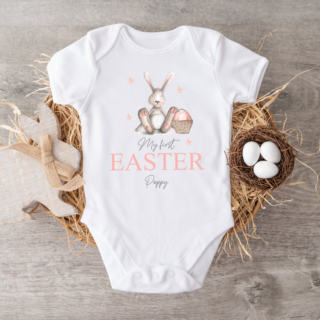 Personalised white Easter baby vest that says 'My First Easter Poppy'. This design features a grey rabbit with pink details sitting next to an Easter basket full with eggs