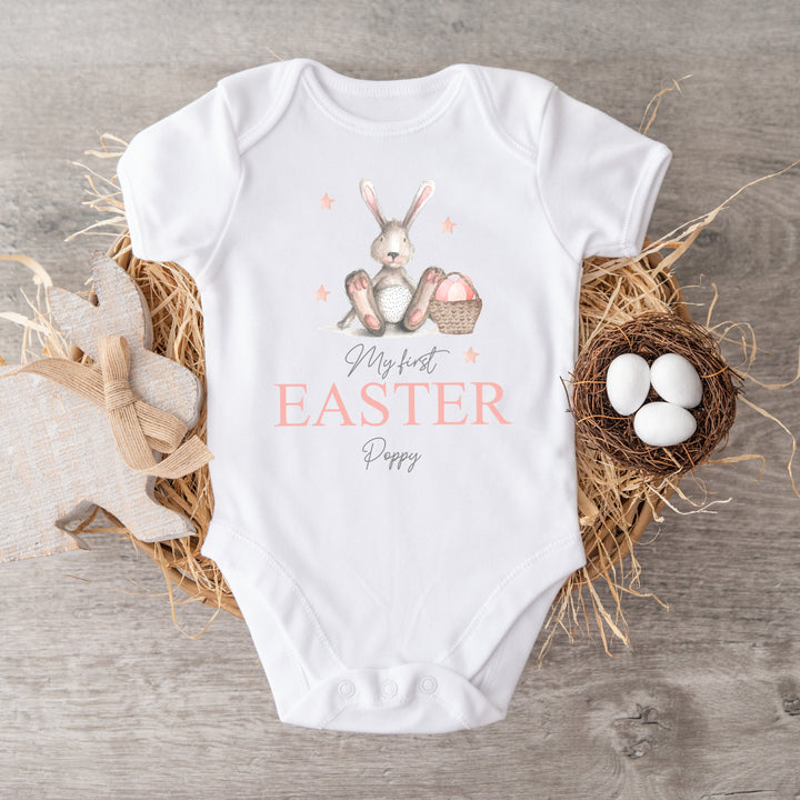 Personalised white Easter baby vest that says 'My First Easter Poppy'. This design features a grey rabbit with pink details sitting next to an Easter basket full with eggs