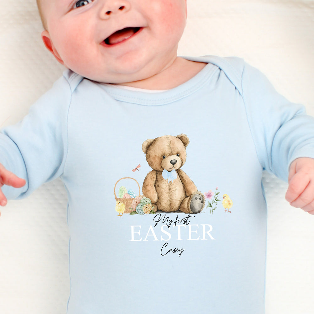 Personalised blue Easter baby grow/sleepsuit that says 'My First Easter Casey'. This design features a teddy bear wearing a blue bow tie sitting next to an Easter basket and 2 chicks