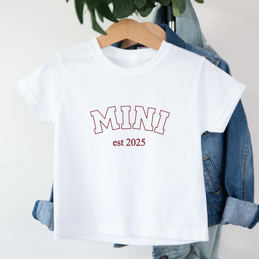 White t-shirt with 'MINI est 2025' text on a hanger with denim jacket.