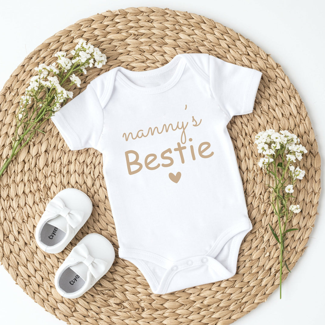 White baby onesie with 'nanny's Bestie' text on a woven mat with white shoes and flowers.