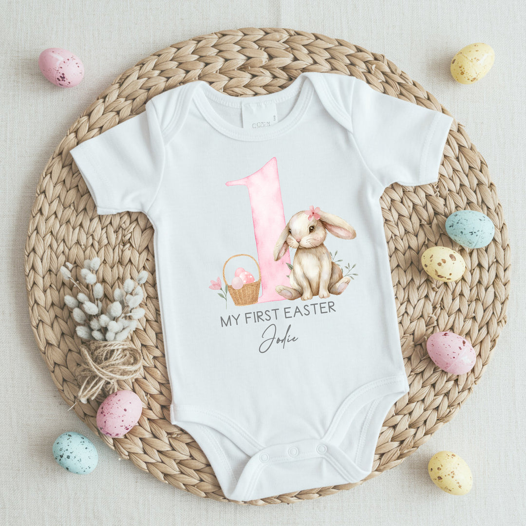 Personalised white Easter baby vest that says 'My First Easter Jodie'. This design features a big pink 1 with a brown bunny one side and an Easter basket the other side