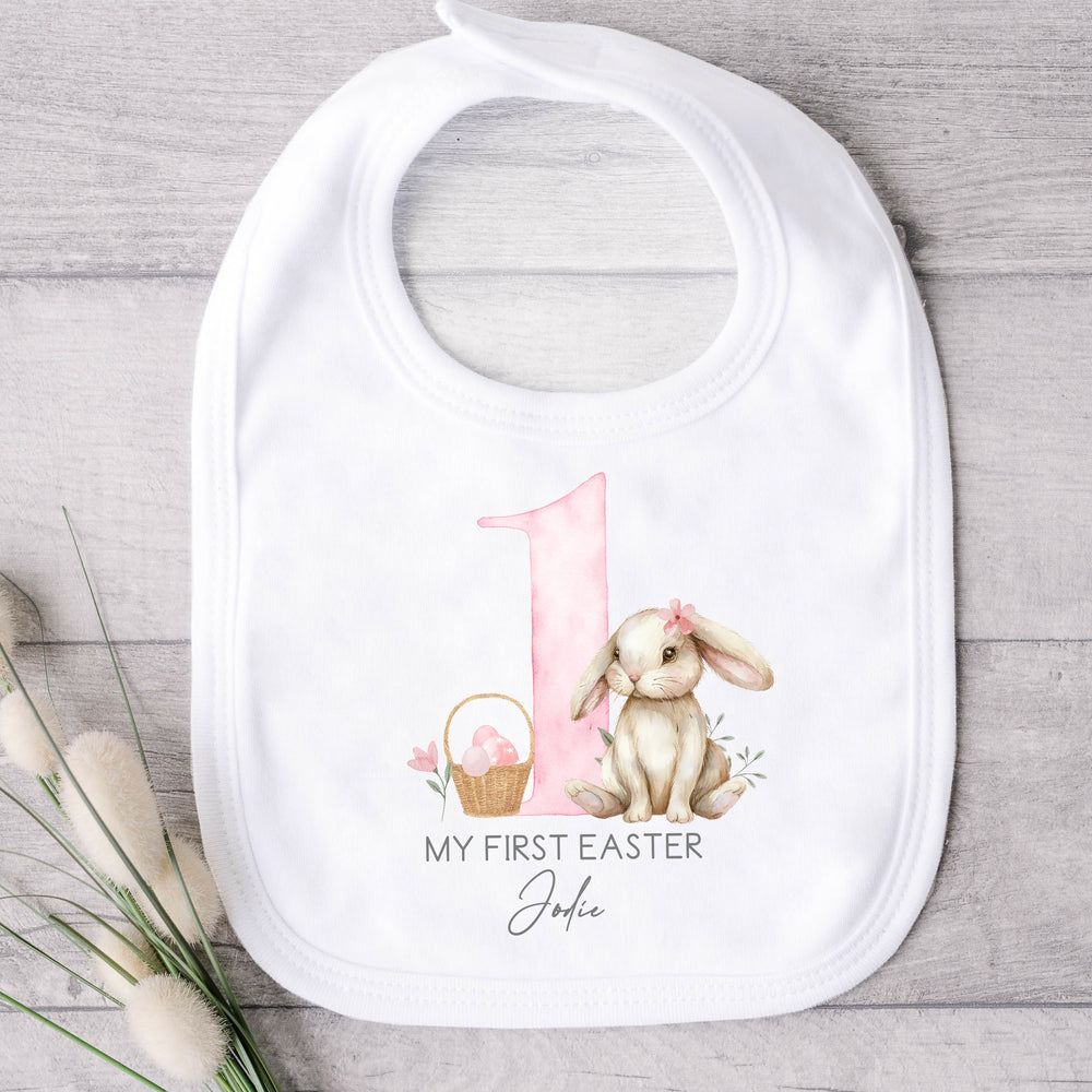 Personalised white Easter baby bib that says 'My First Easter Jodie'. This design features a big pink 1 with a brown bunny one side and an Easter basket the other side