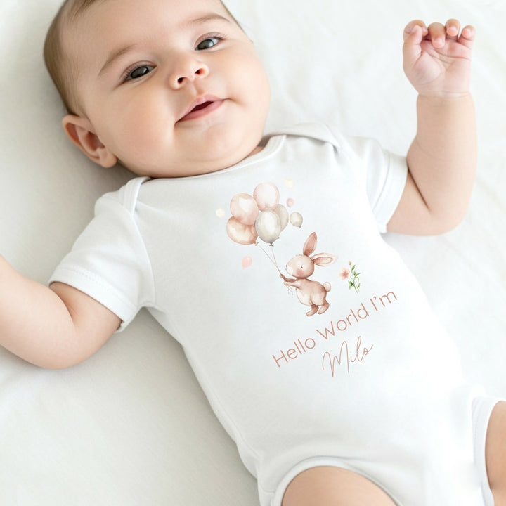 Personalised white baby vest that says 'Hello World I'm Milo'. This design features a light pink rabbit holding onto a bunch of balloons