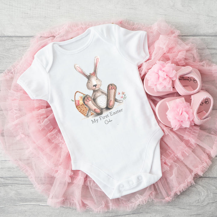 Personalised white Easter baby vest that says 'My First Easter Addison'. This design features a grey bunny with pink details sitting next to an Easter basket