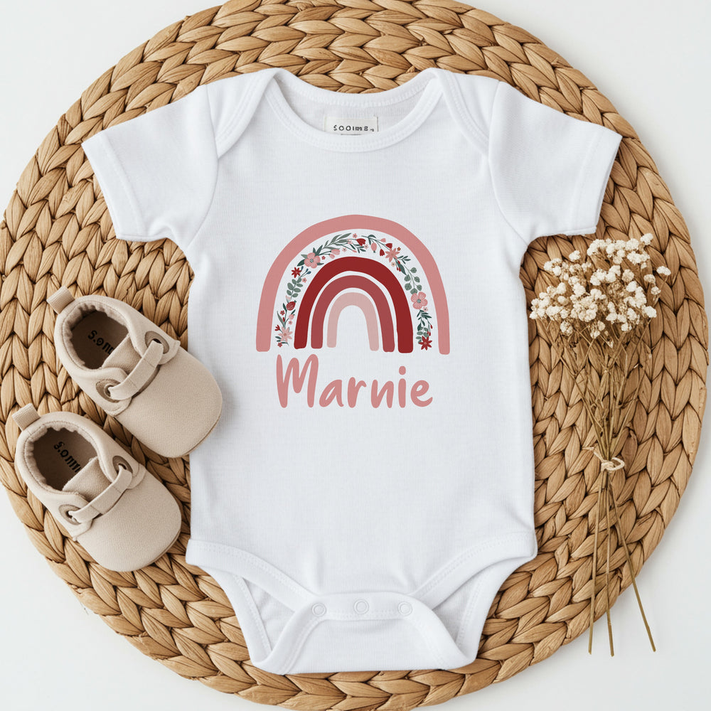 a personalised white baby vest that says 'Marnie'. This design features a pink floral rainbow