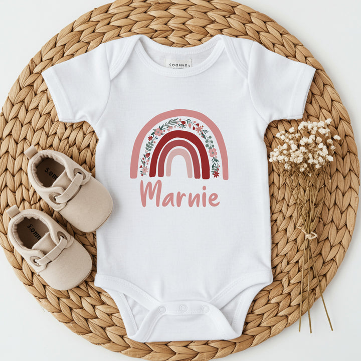 a personalised white baby vest that says 'Marnie'. This design features a pink floral rainbow