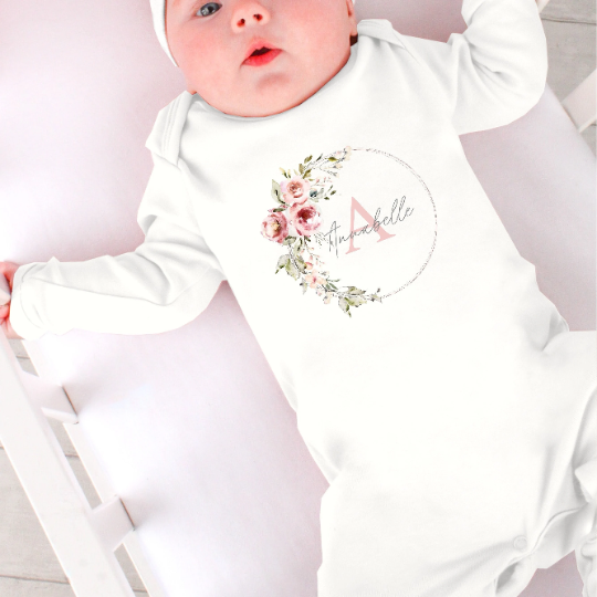 A white babygrow with a floral wreath printed on the chest. In the wreath is printed a name in front of an initial which can be personalised.