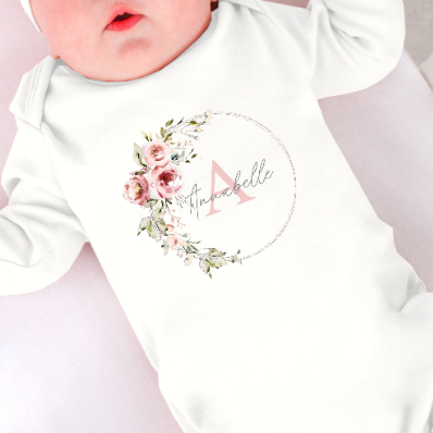 A white babygrow with a floral wreath printed on the chest. In the wreath is printed a name in front of an initial which can be personalised.