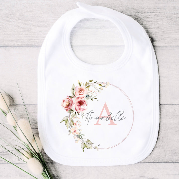 A personalised white baby bib that says 'Annabelle'. This design features a pink floral wreath with a name and initial in the middle
