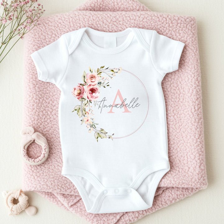 A personalised white baby vest that says 'Annabelle'. This design features a pink floral wreath with a name and initial in the middle