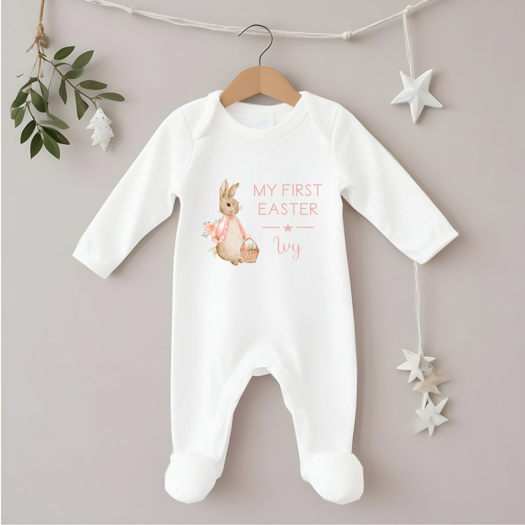 Personalised white Easter baby grow/sleepsuit that says 'My First Easter Ivy'. This design features a rabbit wearing a pink jacket holding an Easter basket full of eggs