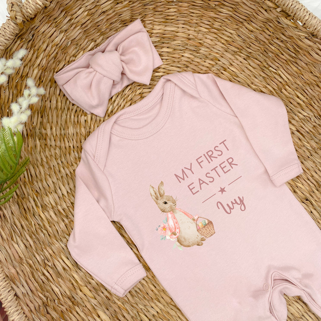 Personalised pink Easter baby grow/sleepsuit that says 'My First Easter Ivy'. This design features a rabbit wearing a pink jacket holding an Easter basket full of eggs