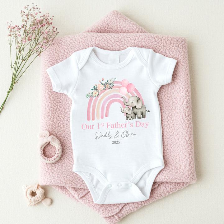 Personalised white Father's Day baby vest that says 'Our 1st Father's Day Daddy & Olivia 2025'. This design features a pink rainbow with 2 grey elephants holding 3 pink balloons