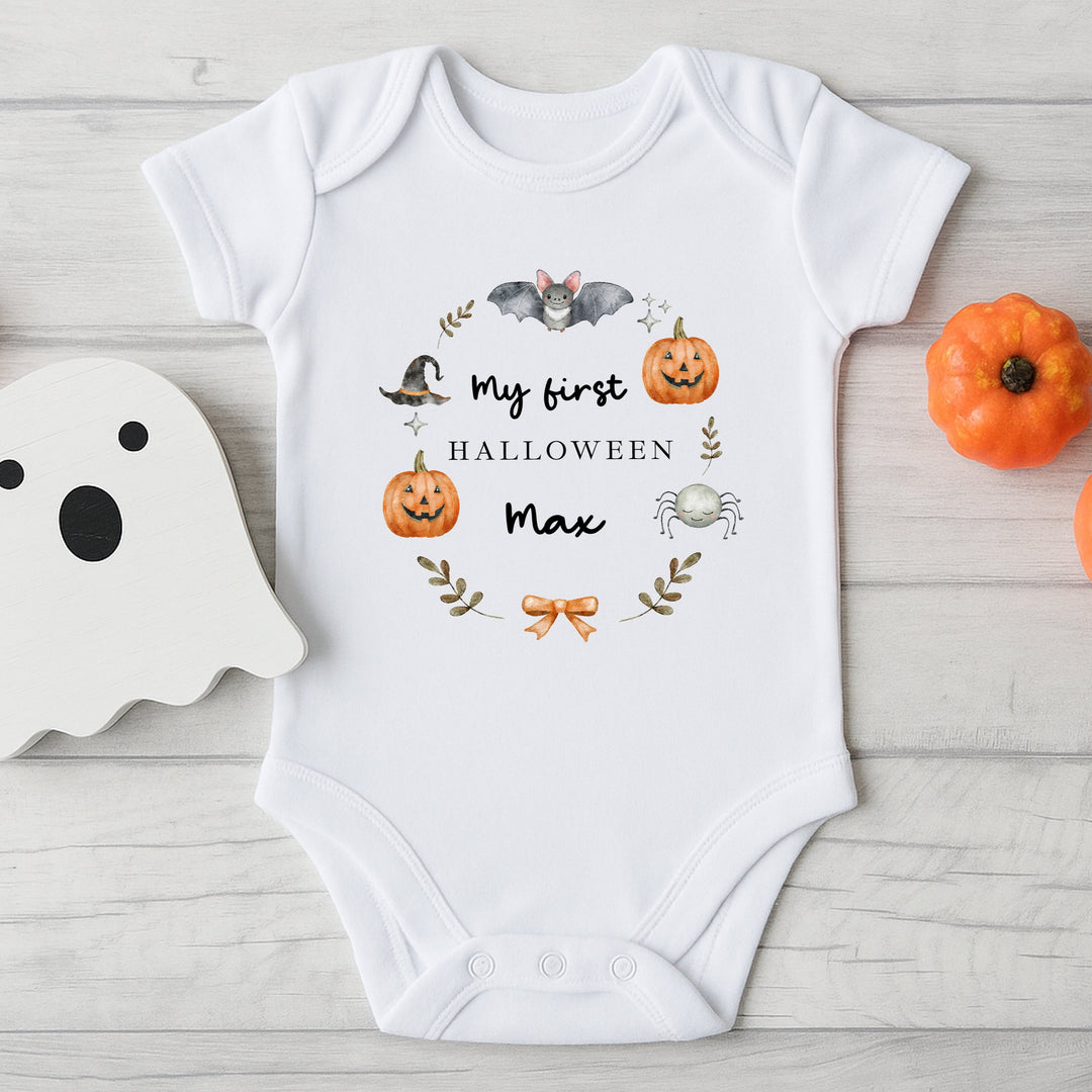 White baby onesie with Halloween-themed design and text on a wooden surface with decorative items.
