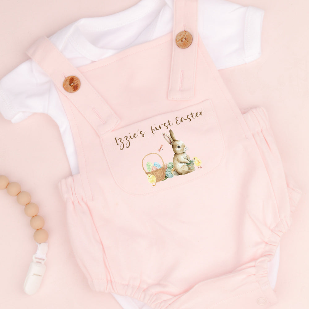 Personalised pink Easter dungarees that say 'Izzie's First Easter'. This design features a brown bunny sittinhg next to an Easter basket full of eggs