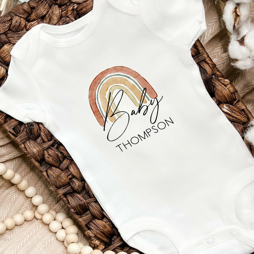 A white baby vest printed with a rainbow in warm neutral colours. The design can be personalised with a choice of name.