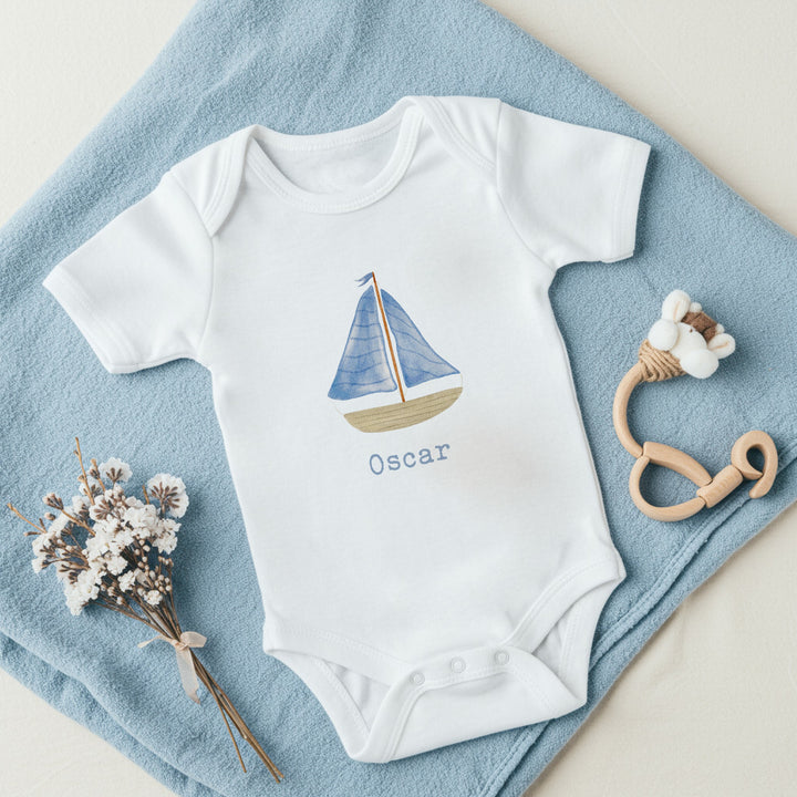 a personalised white baby vest that says 'Oscar'. This design features a blue sail boat