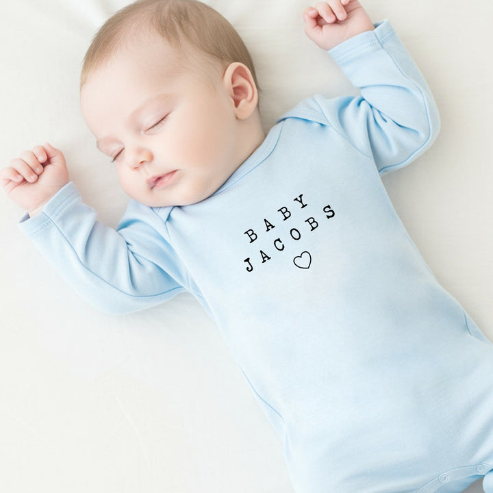 a personalised blue baby grow that says 'Baby Jacobs'. This design features a small heart under the text